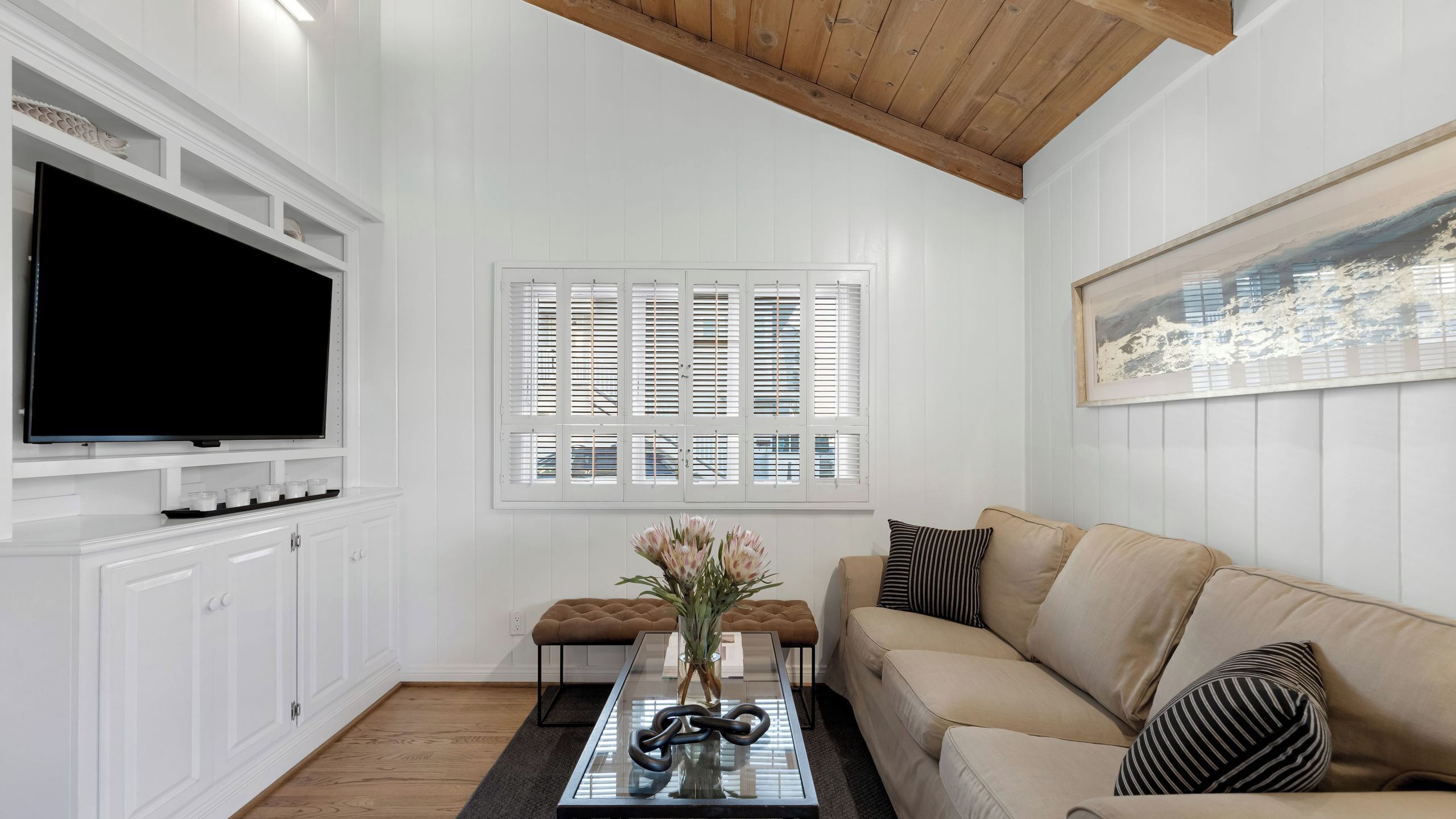 A living room with a flat-screen TV, white paneled walls, a large window with shutters, a beige sofa, and a coffee table with a vase.