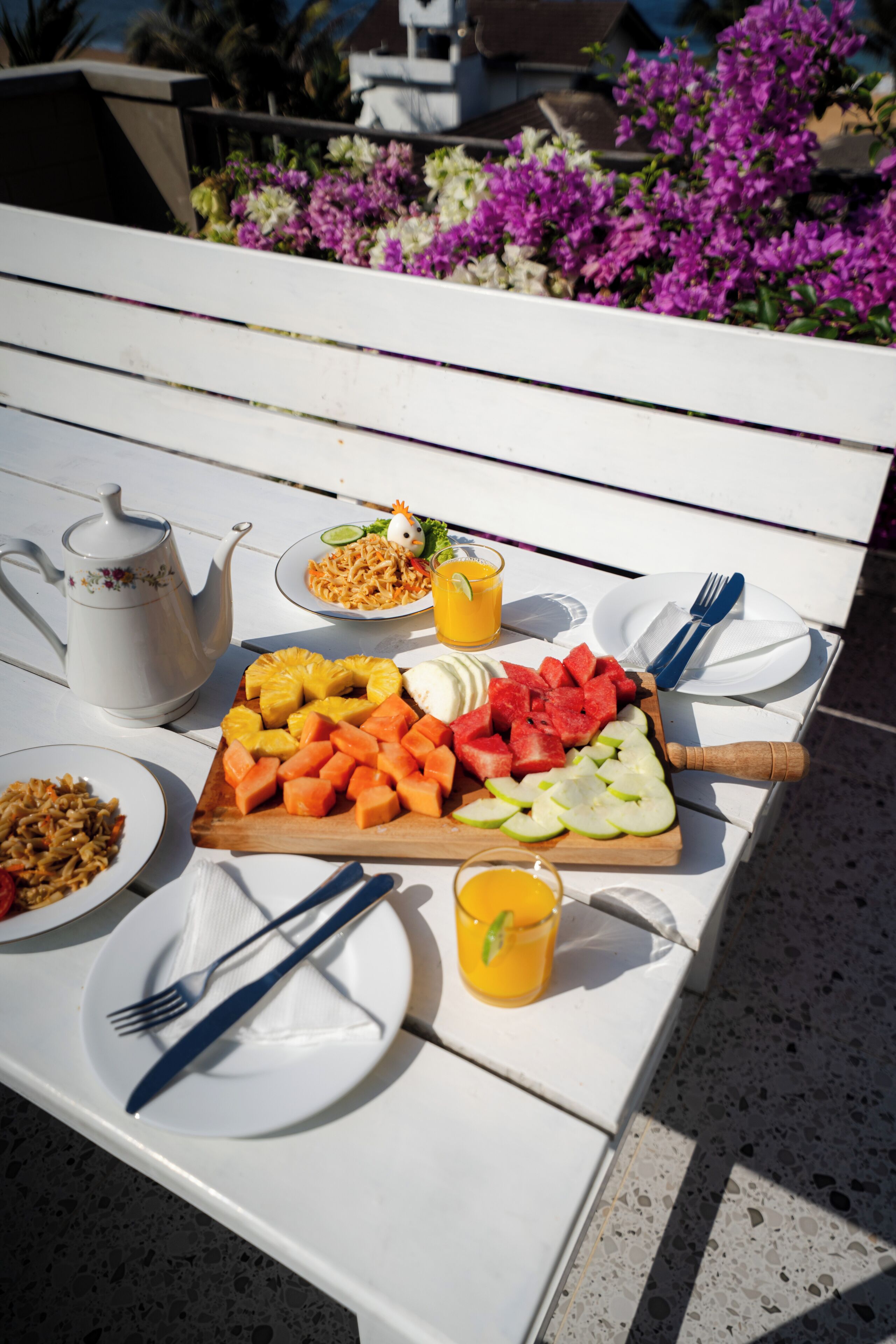 Een tafel gedekt met verschillende soorten eten, waaronder fruit, een drankje en een bord met noedels, wat duidt op een maaltijd in de open lucht.