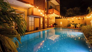 Une piscine illuminée de guirlandes lumineuses, un bâtiment en briques et un balcon.