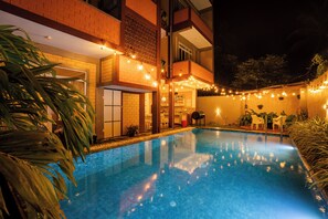Une piscine illuminée de guirlandes lumineuses, un bâtiment en briques et un balcon.