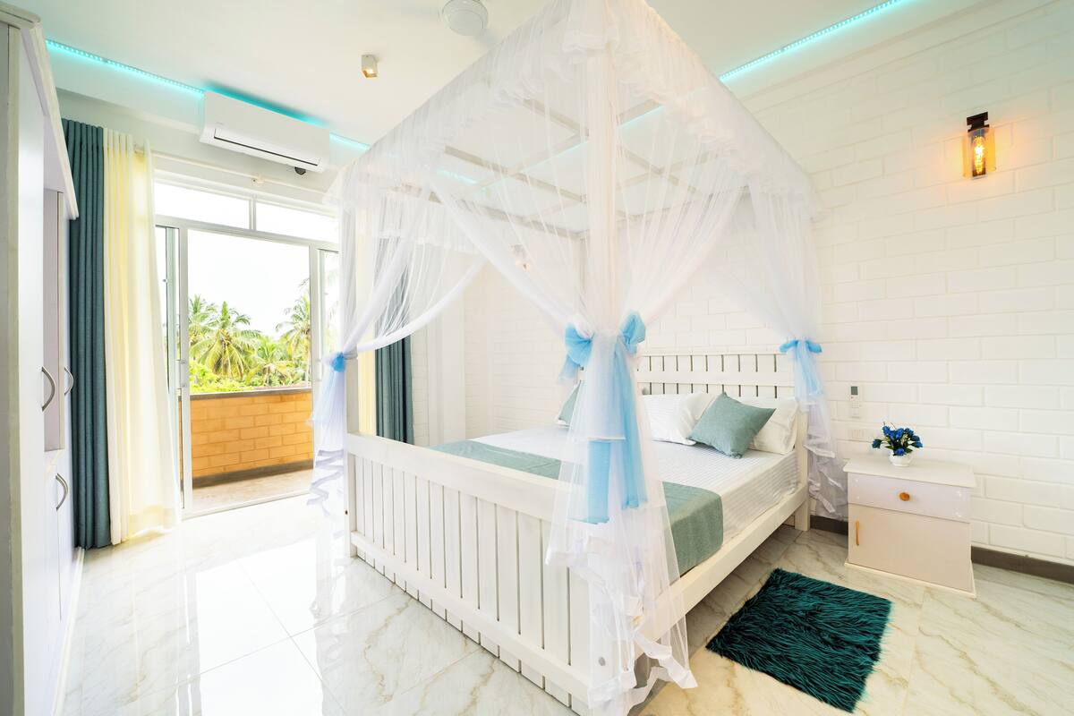 Une chambre à coucher avec un lit à baldaquin blanc, une table de chevet avec une lampe et une vue sur la verdure à l’extérieur.