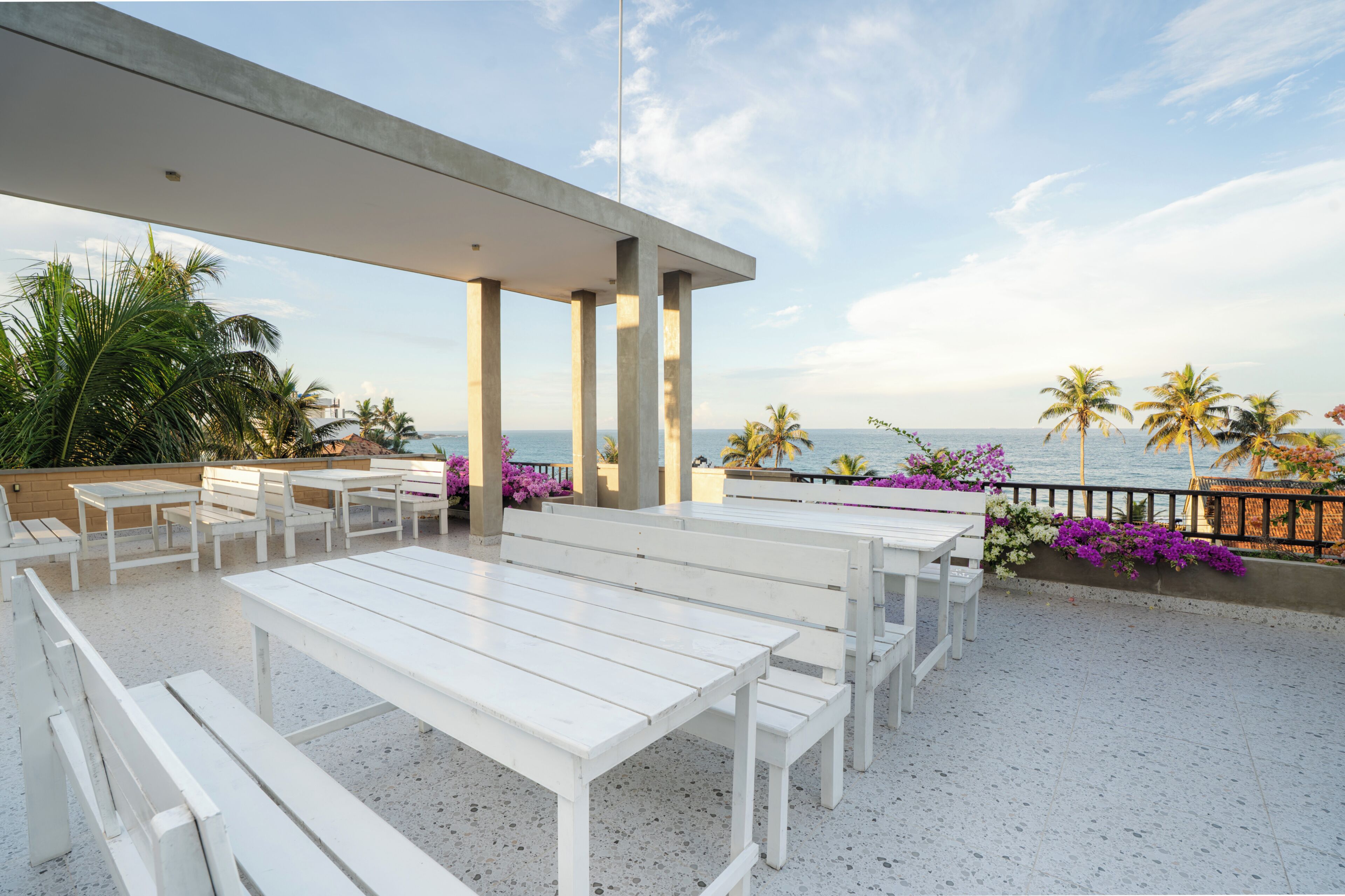 An outdoor seating area with white tables and benches, a concrete ceiling, and a view of the ocean with palm trees.