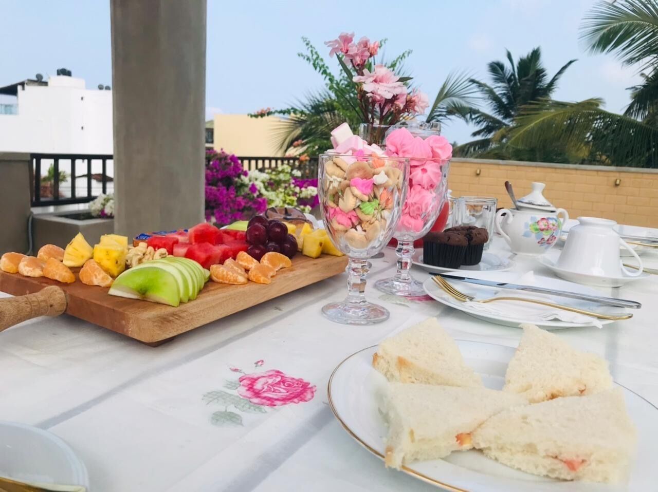 Una mesa dispuesta con diversas opciones de comida, entre ellas frutas, sándwiches y postres, con vistas a palmeras y edificios al fondo.