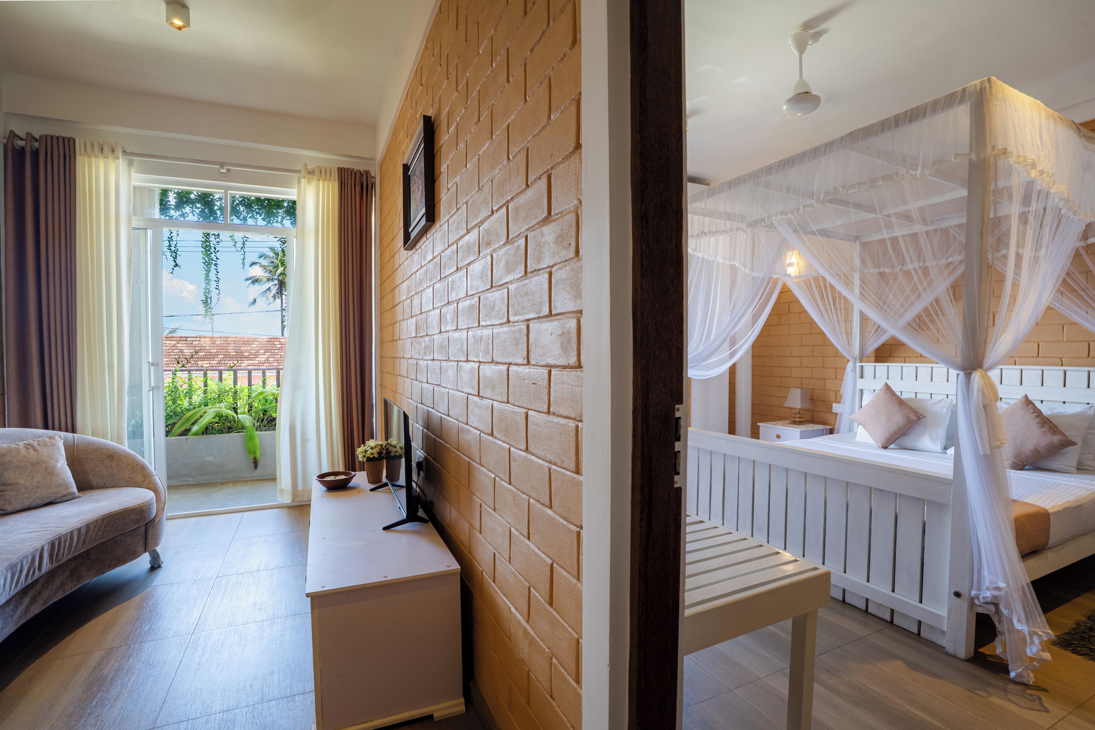 A room with a brick wall, a bed with a canopy, a sofa, a small table, and a view of the outdoors.