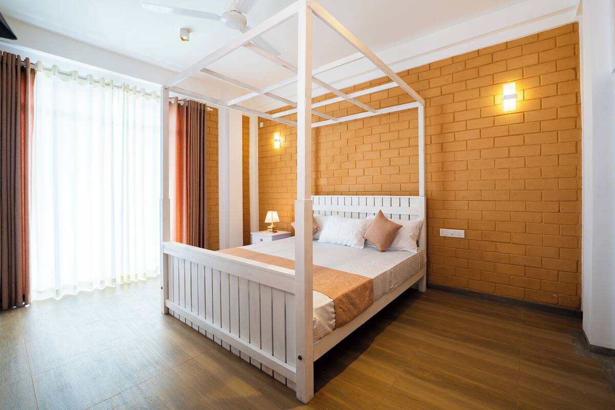 Un lit à baldaquin avec un cadre blanc, un plancher en bois et un mur en briques agrémenté d’un luminaire.