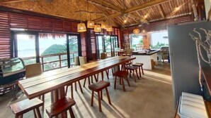 A spacious dining area with a long wooden table and benches, large windows offering a view of the outdoors, and a ceiling with exposed wooden beams. - 89 Villas Resort and Spa by Hiverooms (El Nido)