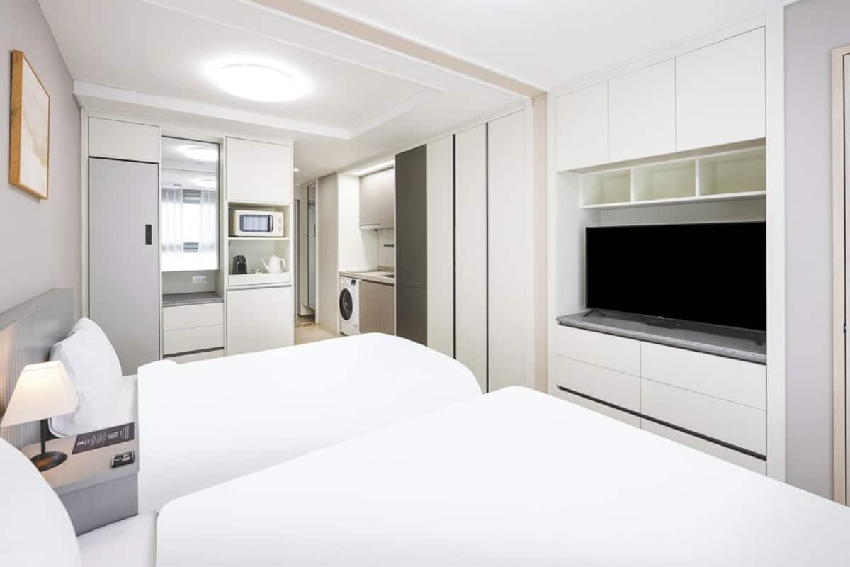 Ein Hotelzimmer mit Bett, Fernseher, Schreibtisch mit Lampe und einer Küchenzeile im Hintergrund.
