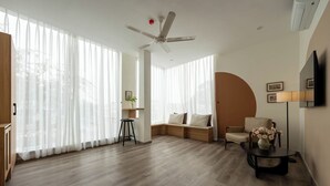 Un salon spacieux doté d’un plancher en bois, d’un ventilateur de plafond, d’un canapé, d’un fauteuil, d’une petite table avec un vase et d’un téléviseur fixé au mur.