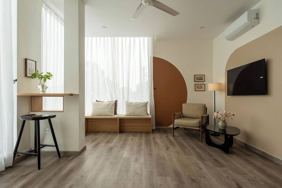 A modern living room with a wooden floor, a sofa with cushions, a chair, a small table with a vase, and a mounted TV.