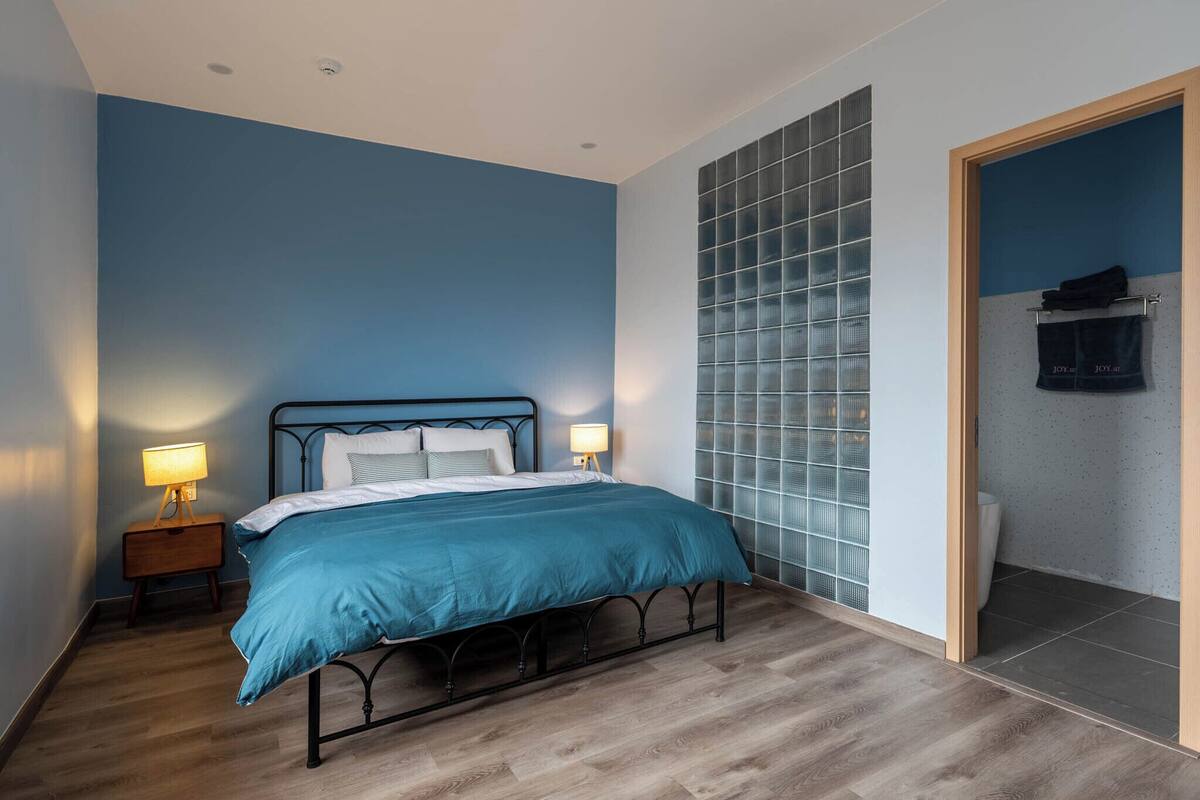 A bedroom with a bed, two bedside lamps, a nightstand, and a wall with a window.