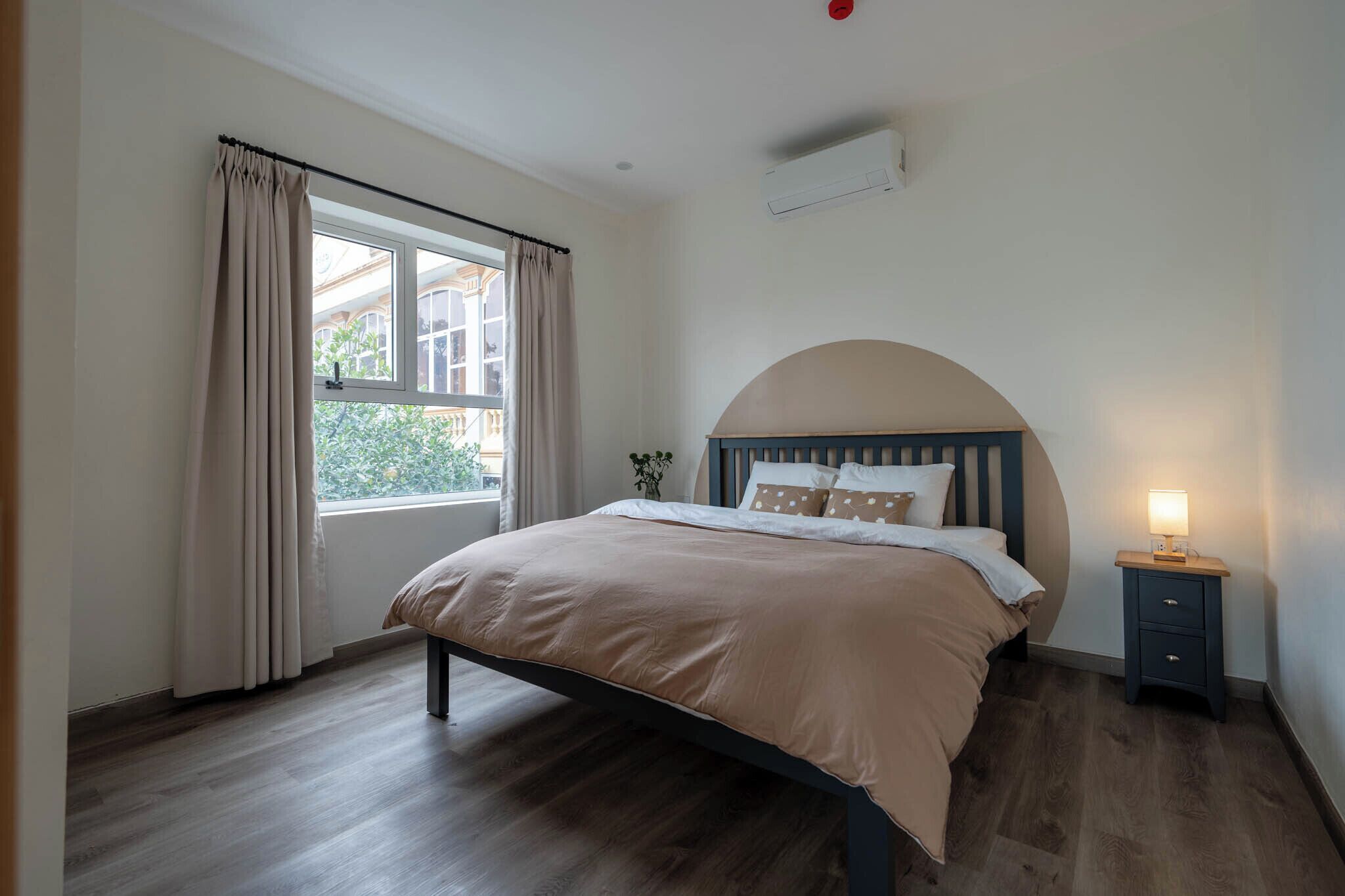 A bedroom with a bed, a nightstand, a window with curtains, and an air conditioning unit.
