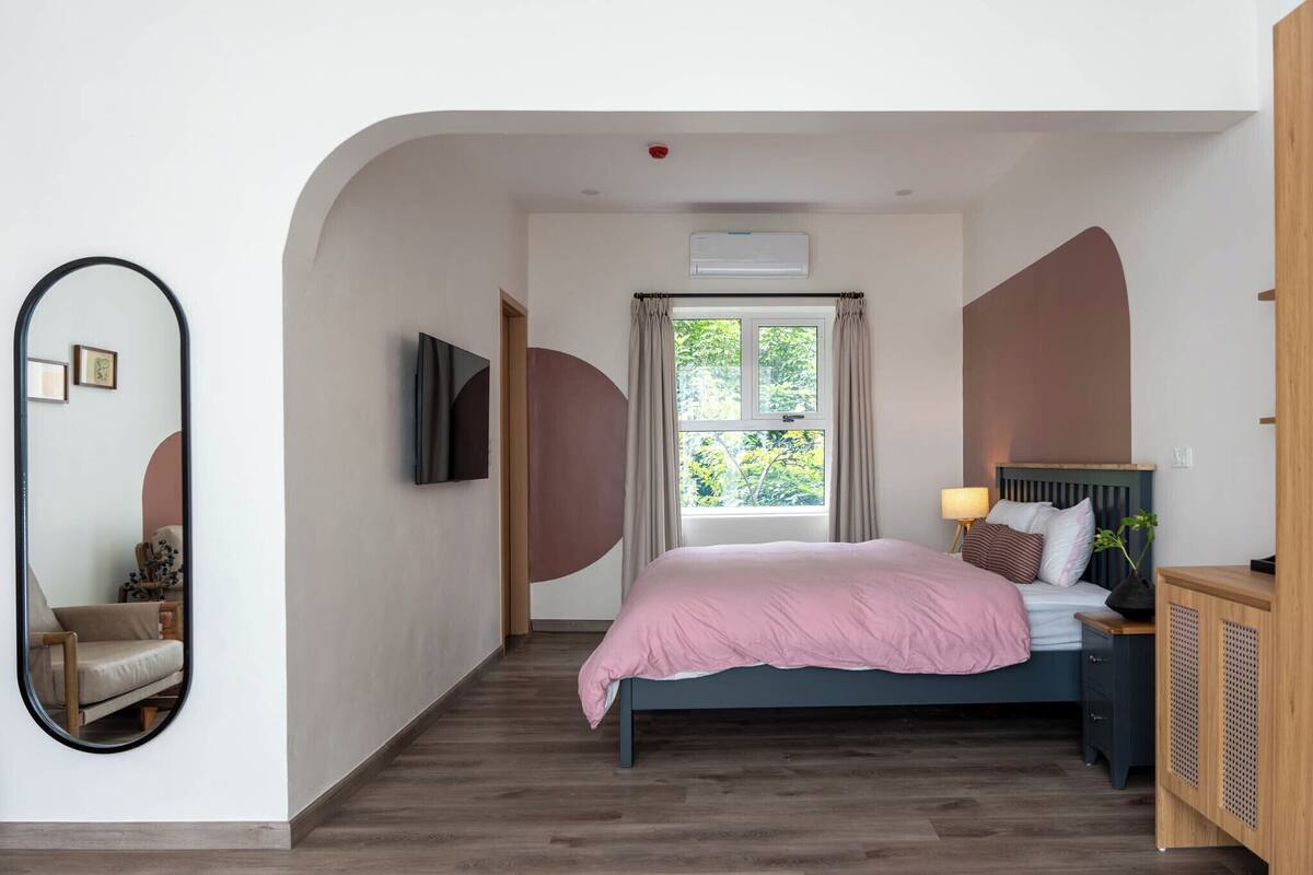 A bedroom with a bed, a mirror, a window with curtains, a bedside table with a lamp, and a TV mounted on the wall.