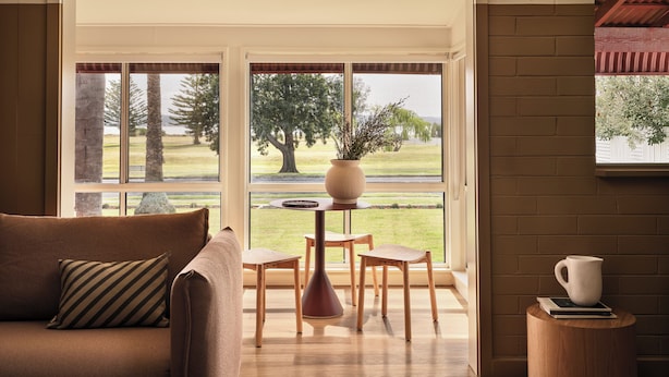 A cozy living room with a sofa, a coffee table, and a view of a grassy area through large windows. - Casita Motel (Batehaven)