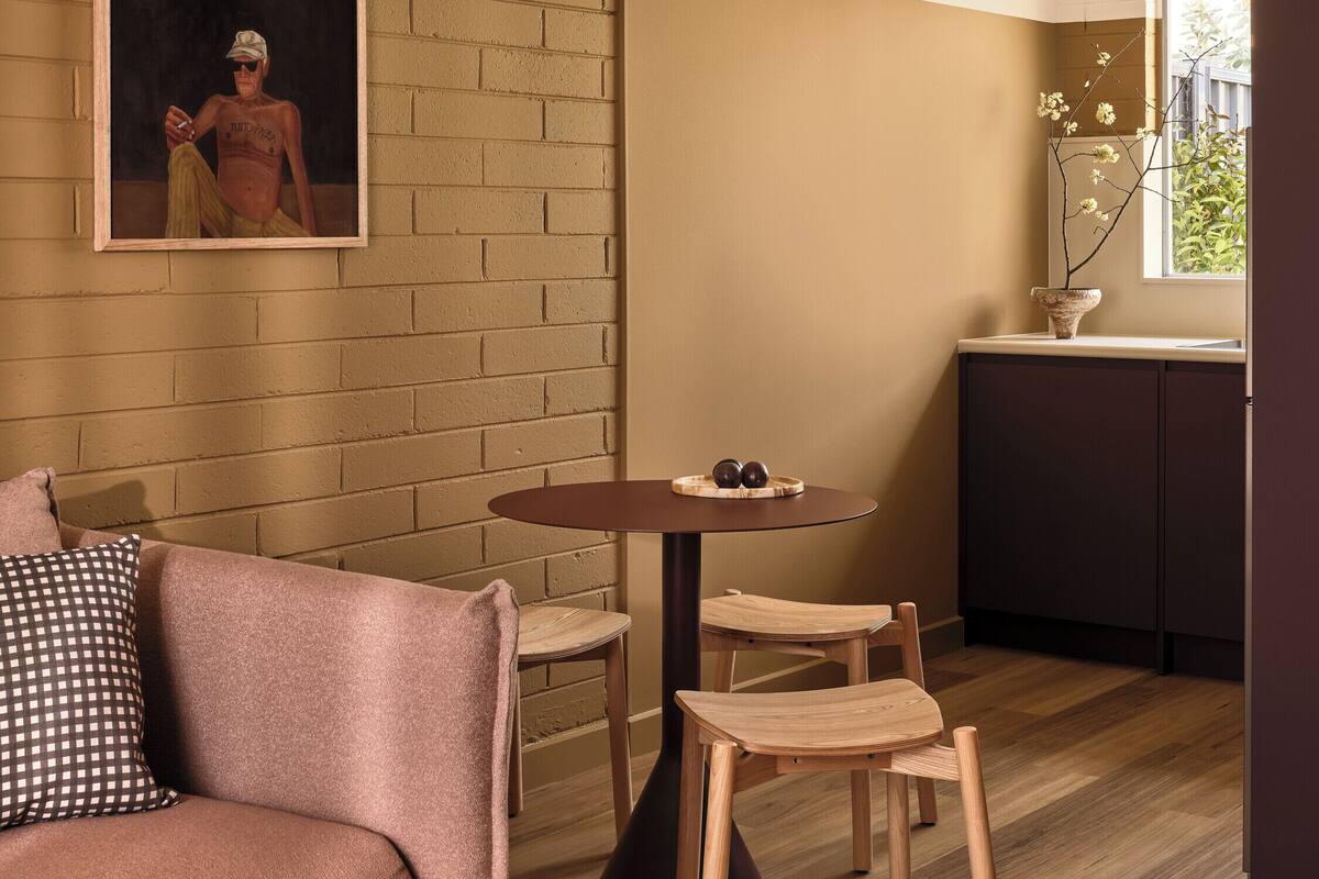 A cozy living room with a brick wall, a painting, a round table with chairs, a sofa, and a small kitchen area with a window.