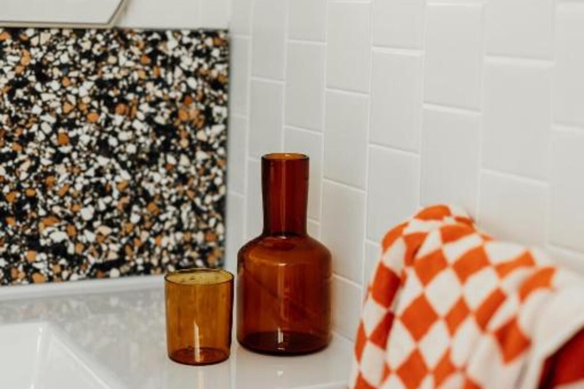 A bathroom with white tiled walls, a decorative wall piece, a wooden bath mat, and a small wooden container.