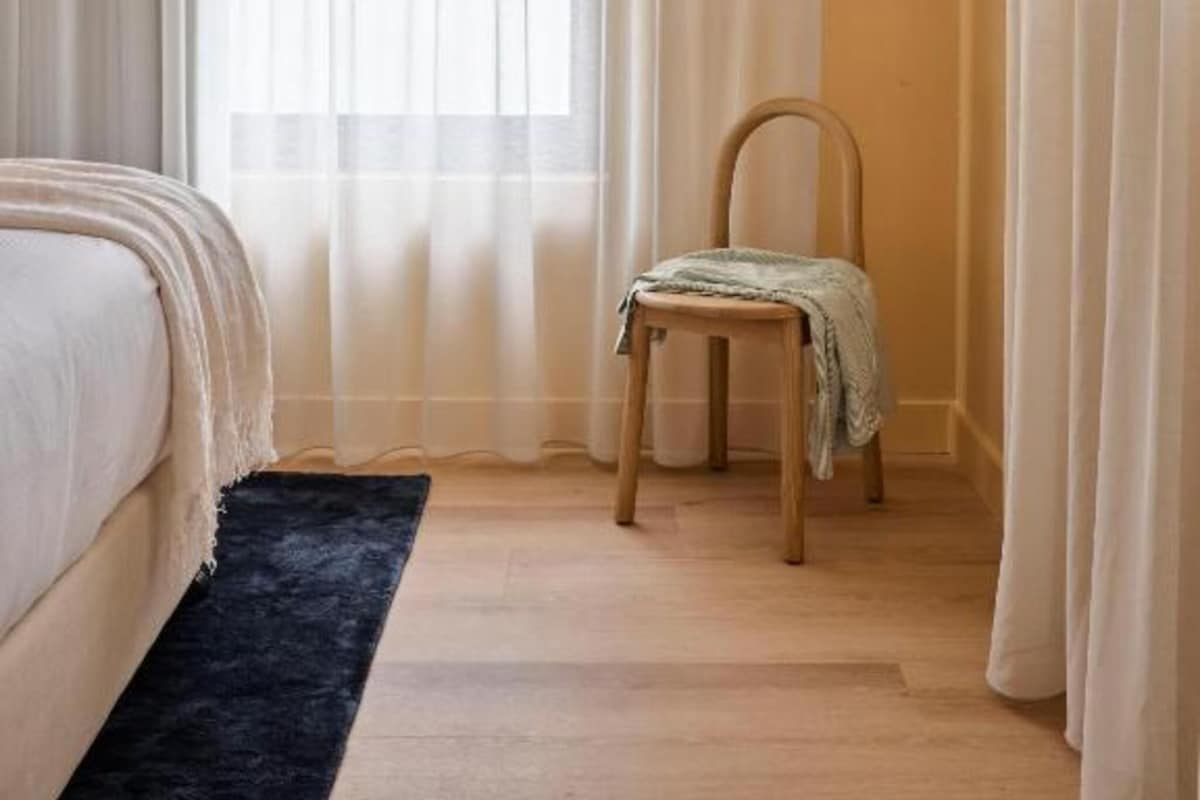A bedroom with a bed, a chair, a window with sheer curtains, and an air conditioning unit.