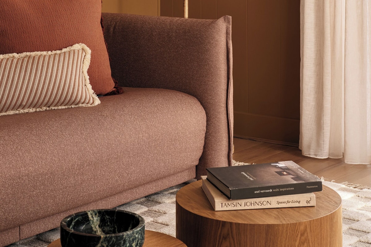 A brown sofa with striped pillows, a round wooden side table with a book and a bowl, and a floor lamp.