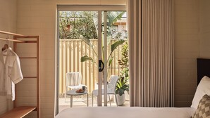 A bedroom with a bed, a wooden shelf, a chair, a table with a plate and a cup, and a view of a balcony with a plant and a fence. - Casita Motel (Batehaven)