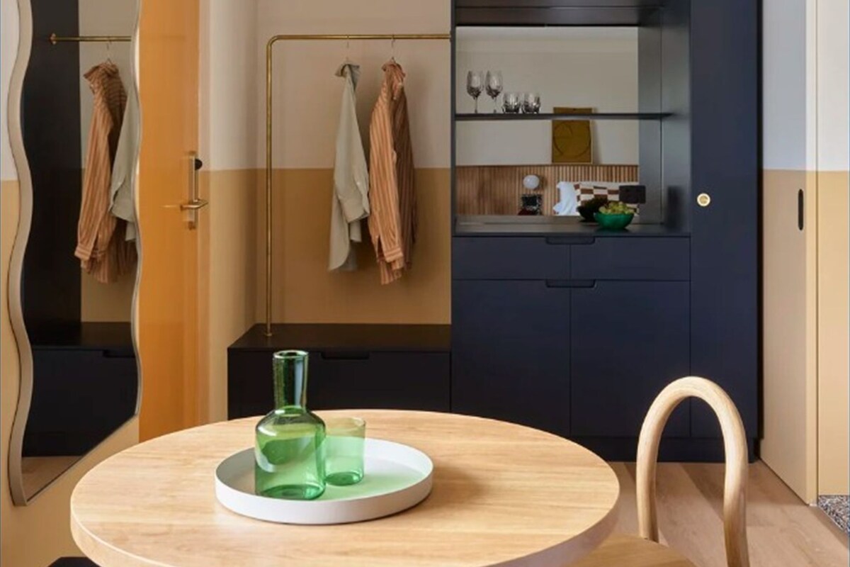 A compact room with a round wooden table, a chair, a mirror, and a dark blue cabinet with a kitchen counter visible in the background.