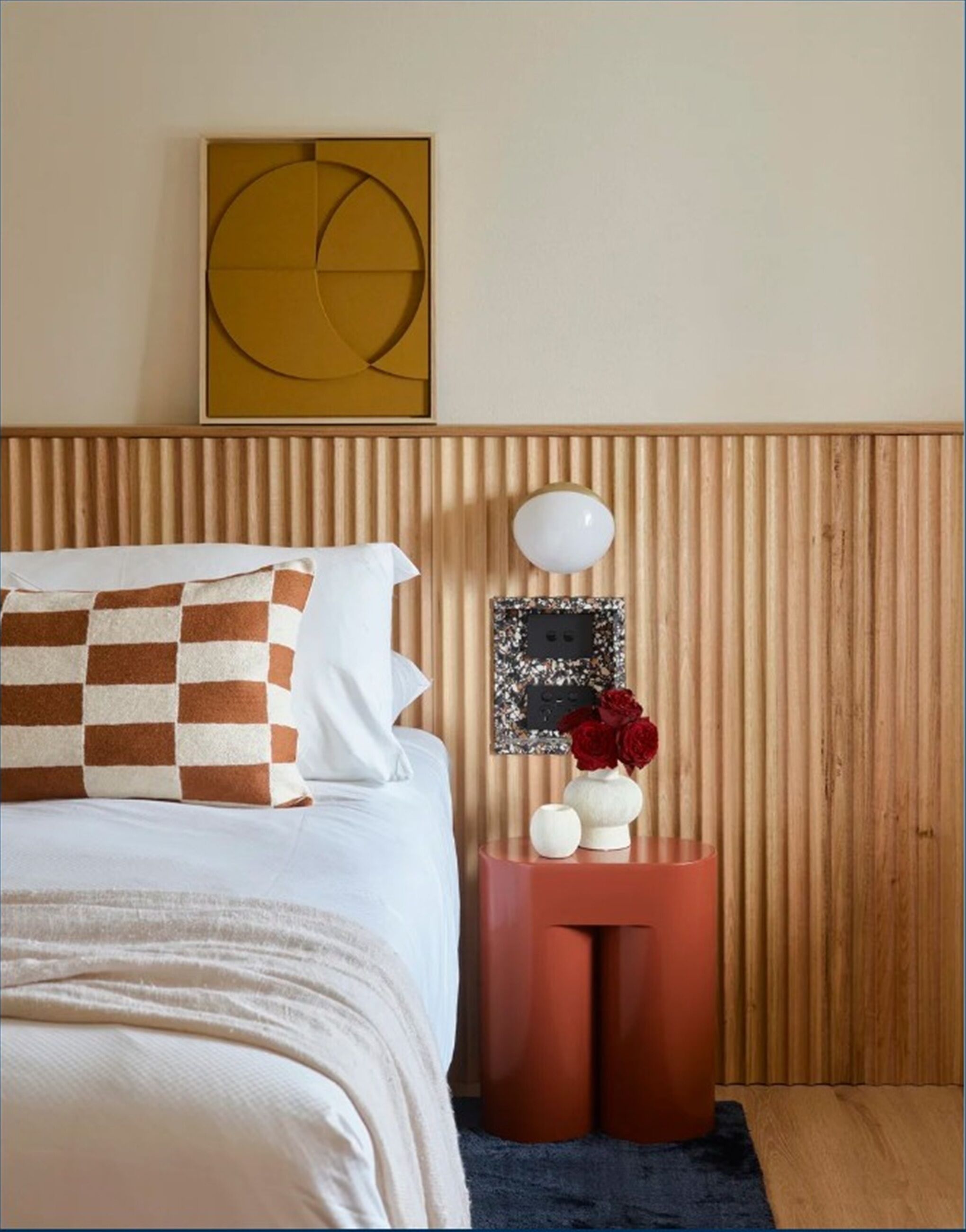 A bedroom with a bed, a bedside table with a vase of flowers, a wall-mounted picture, and a wooden headboard.