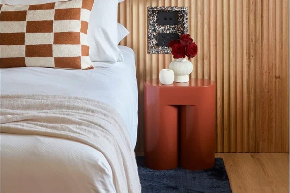A bedroom with a bed, a bedside table with a vase of flowers, a wall-mounted picture, and a wooden headboard.