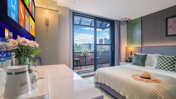 Une chambre d’hôtel avec un lit, un bureau agrémenté d’un vase de fleurs, un téléviseur affichant des icônes colorées et un balcon offrant une vue sur les bâtiments.