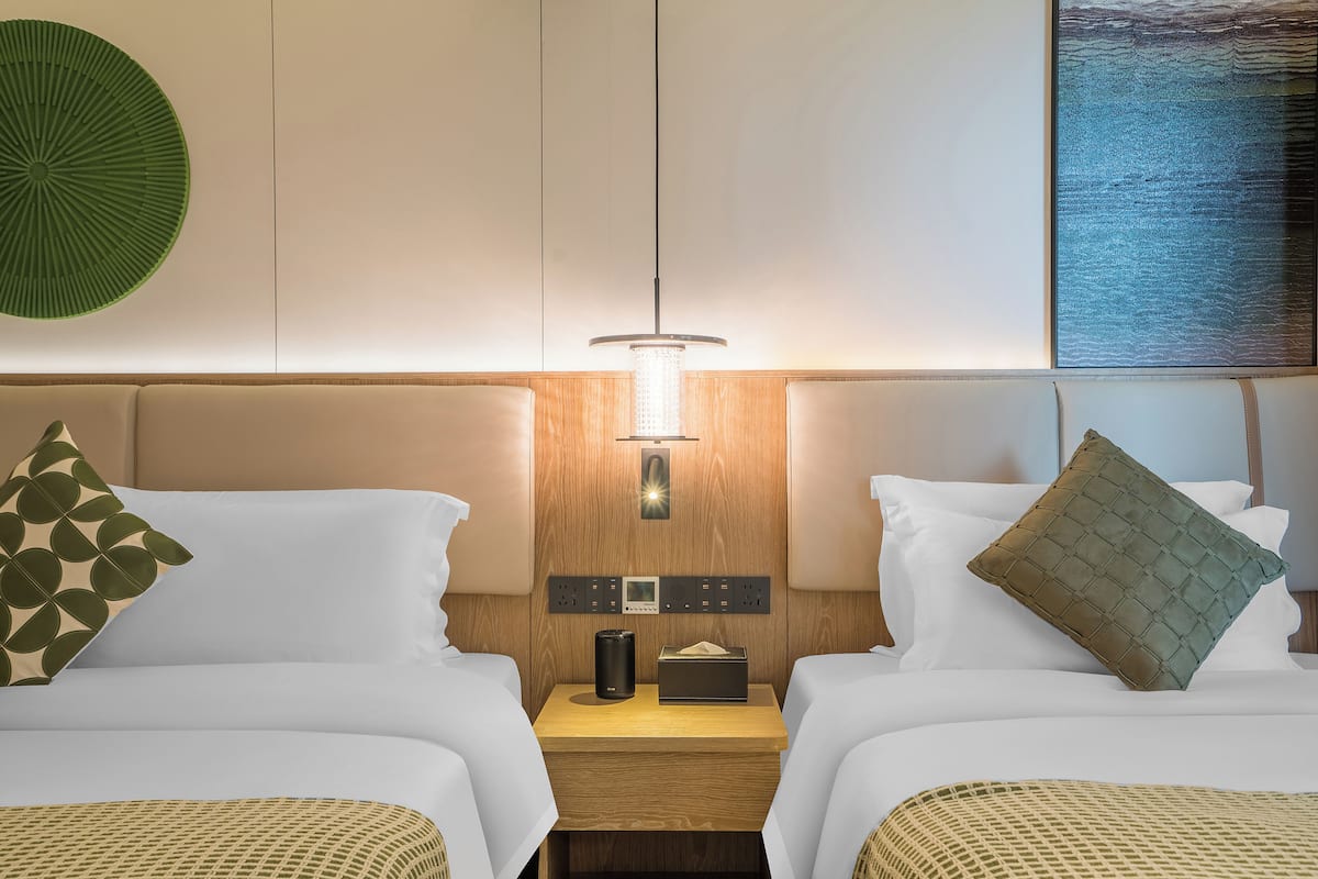 Una habitación de hotel con dos camas, una mesita de noche con lámpara, un panel de control y una obra de arte colgada en la pared.