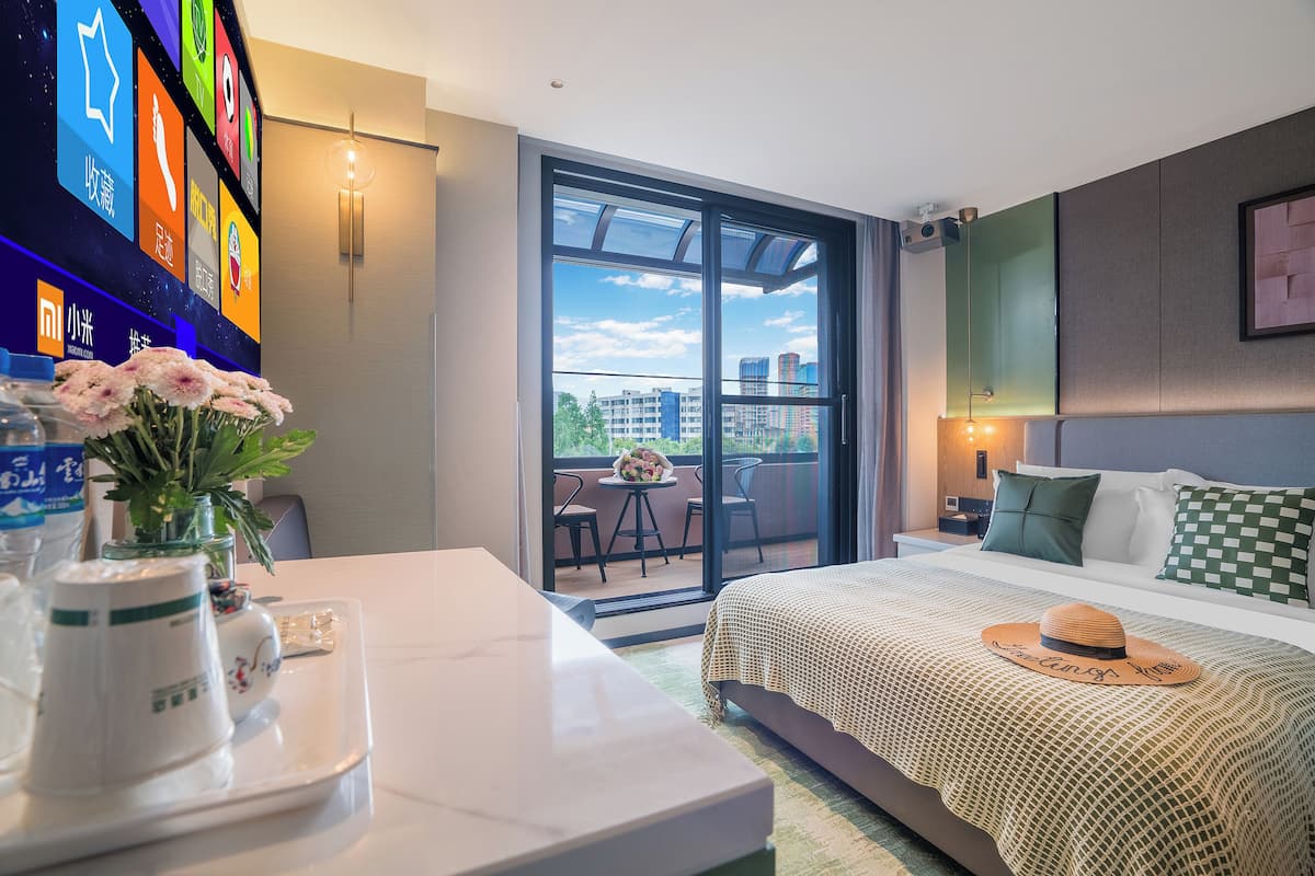 A hotel room with a bed, a desk with a vase of flowers, a TV on the wall, and a balcony with a view of buildings.