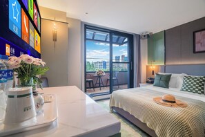 Una habitació d'hotel amb un llit, una taula de treball amb un vas de flors, un televisor penjat a la paret i un balcó amb vista a edificis.