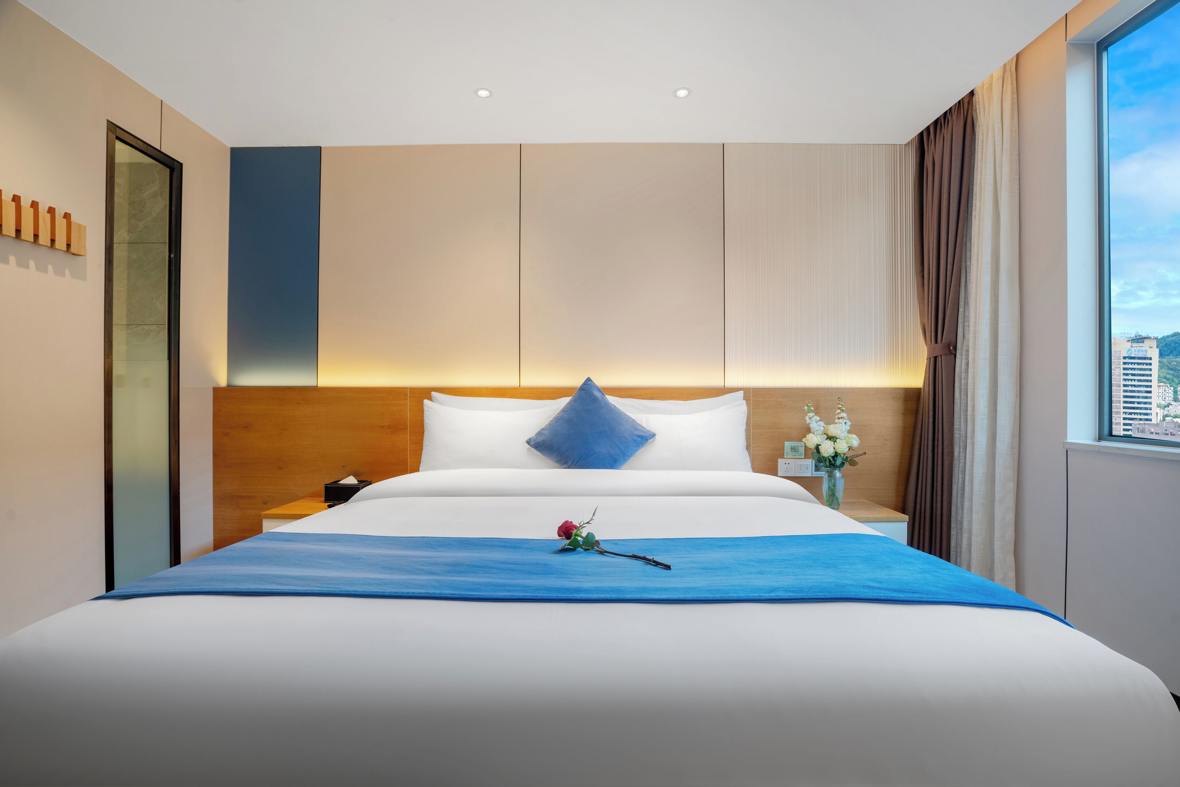 Ein Hotelzimmer mit einem großen Bett, einem Nachttisch, einer Blumenvase und Blick auf die Stadt durch das Fenster.