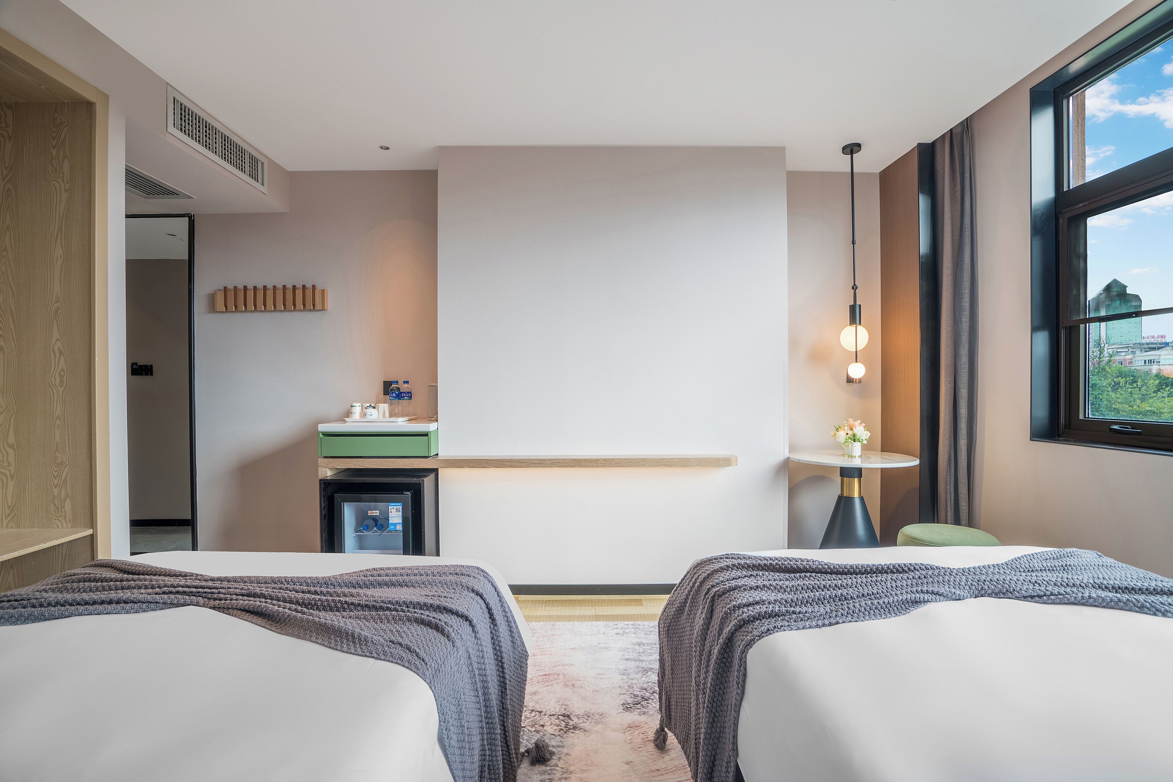 A hotel room with two beds, a small desk, a window with a city view, and a ceiling vent.