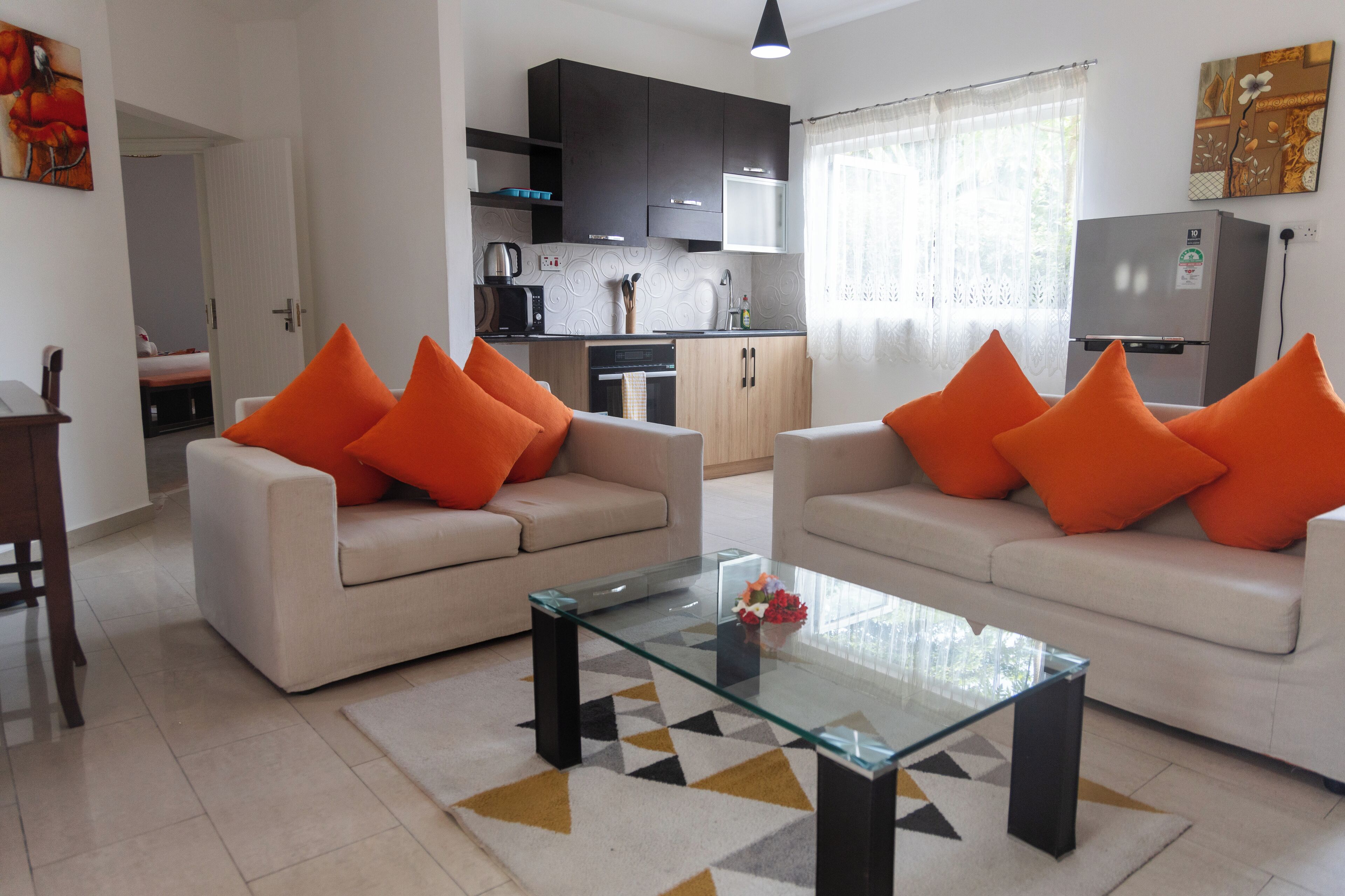 Una sala de estar moderna con dos sofás color beige, cojines naranjas y una mesa de centro de vidrio, con una cocina al fondo.