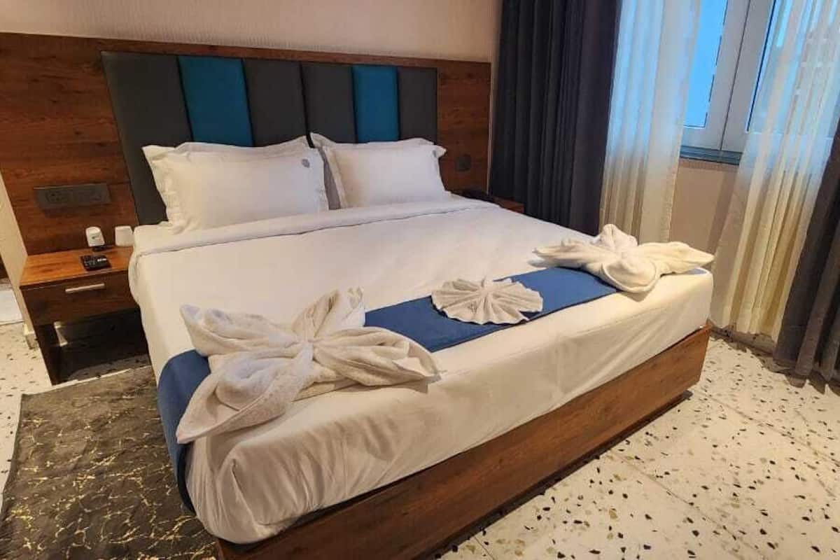 A hotel room with a large bed, wooden headboard, and a blue accent pillow. There is a nightstand with a remote control and a glass of water. The room has a window with curtains and a carpeted floor.