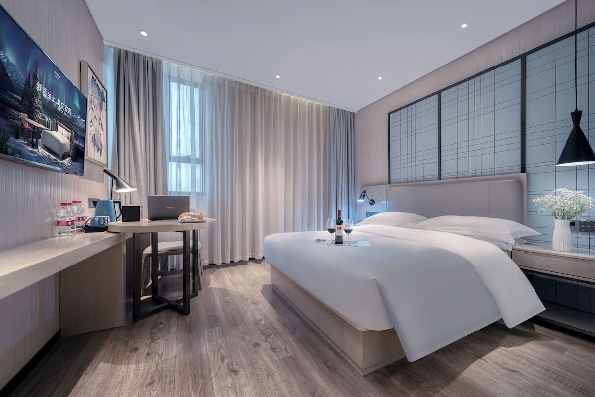Une chambre d’hôtel moderne avec un grand lit, un bureau avec un ordinateur portable, une petite table avec une bouteille et des verres, et une grande fenêtre avec des rideaux.