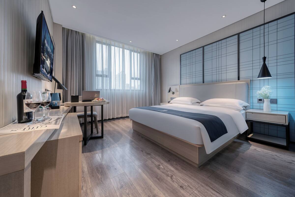 Une chambre d’hôtel moderne dotée d’un grand lit, d’un bureau avec un ordinateur portable, d’un téléviseur fixé au mur et d’une grande fenêtre avec des rideaux.