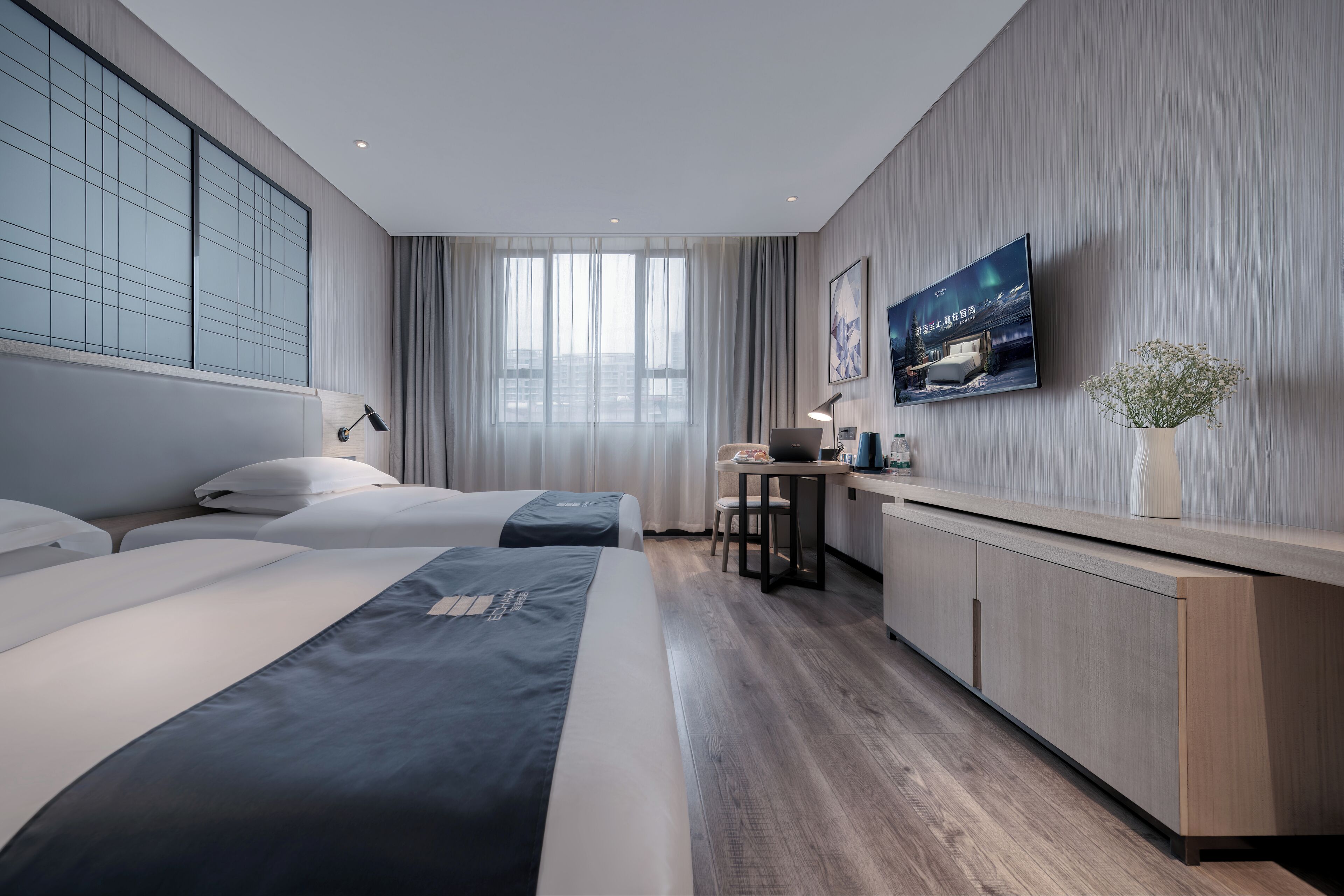 A modern hotel room with two beds, a desk, a chair, and a television on the wall.