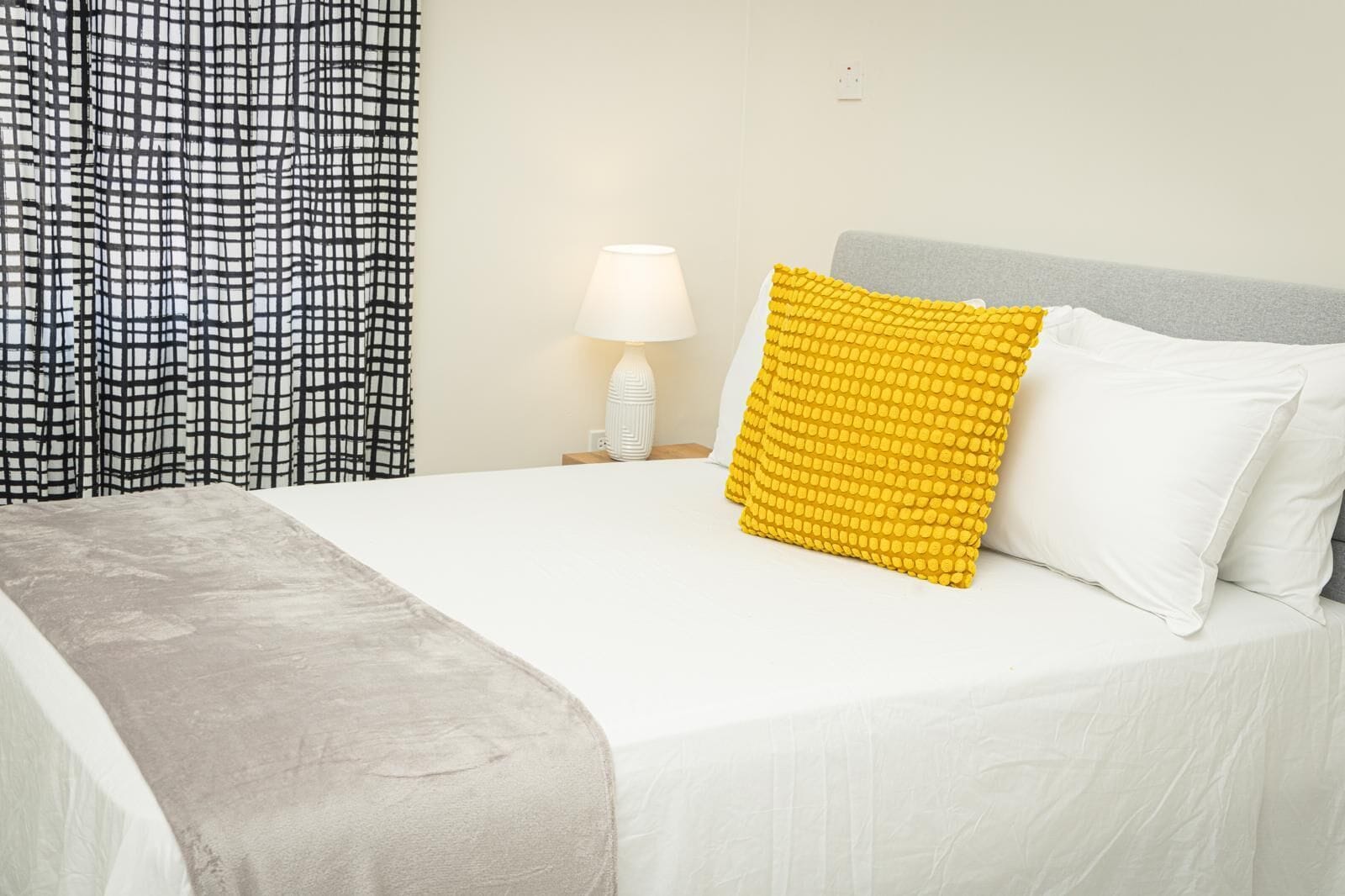 A neatly made bed with white and yellow pillows, a grey blanket, and a white lamp on a bedside table.
