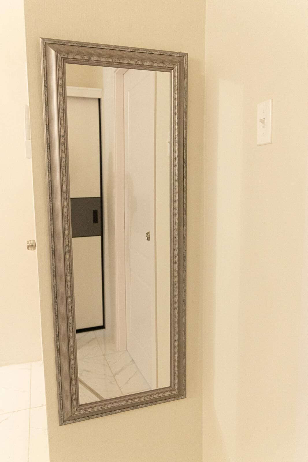 A mirror with a decorative frame reflecting a hallway with a door and a light switch.