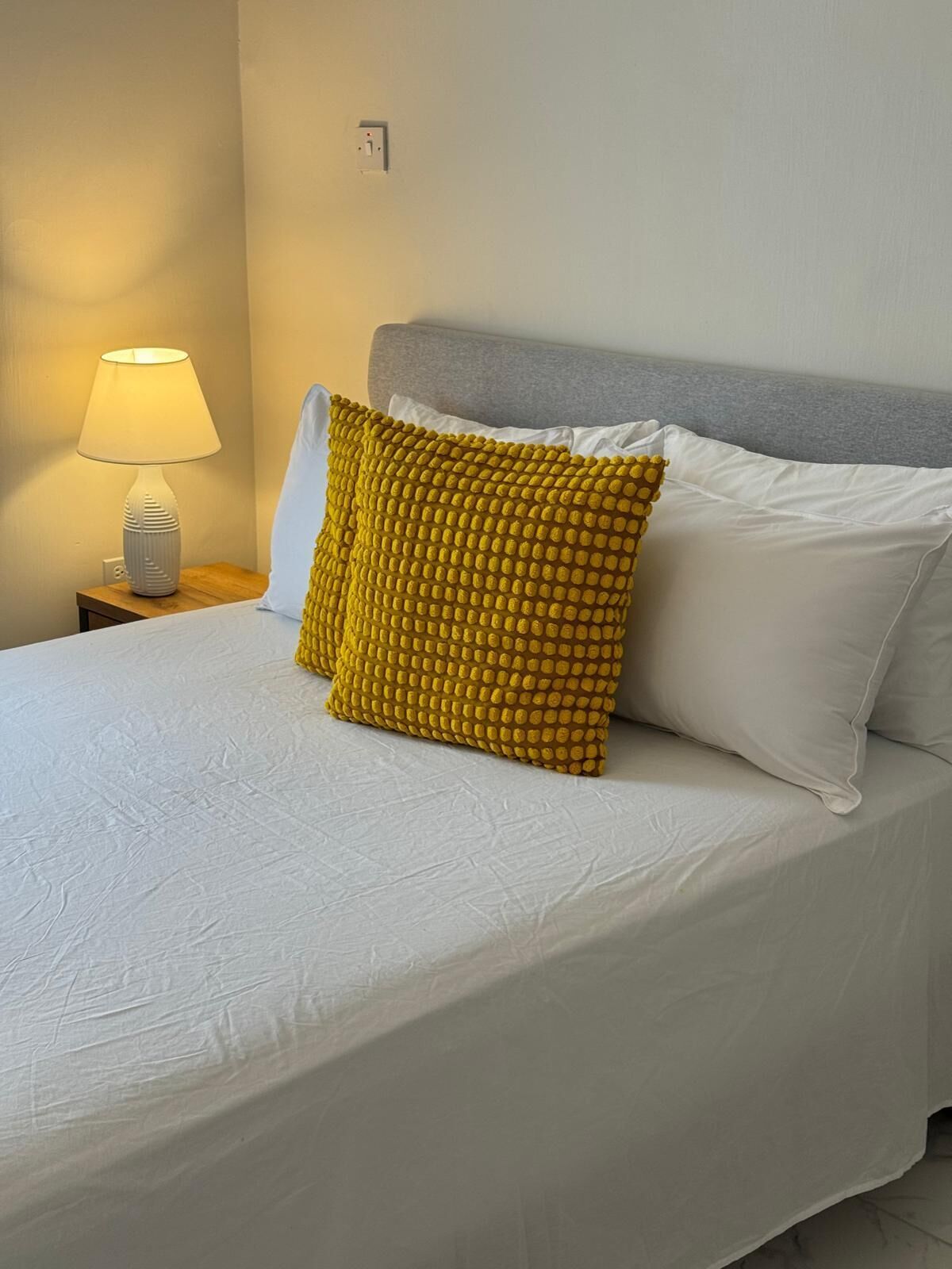 Un lit bien fait, avec une literie blanche, une tête de lit grise et un coussin jaune à texture.