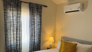 Une chambre d’hôtel avec un lit, une table de chevet avec une lampe, une fenêtre avec des rideaux à motifs et un climatiseur fixé au mur.