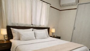 A neatly made bed with white linens and a beige throw blanket, two bedside lamps, a wooden headboard, and an air conditioning unit mounted on the wall.