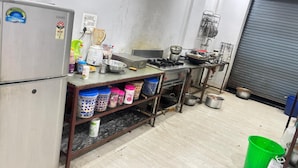 A kitchen with a refrigerator, stove, and various cooking utensils. There are storage containers on shelves and a green bucket on the floor. - Hotel Hari Inn  (Jaipur)