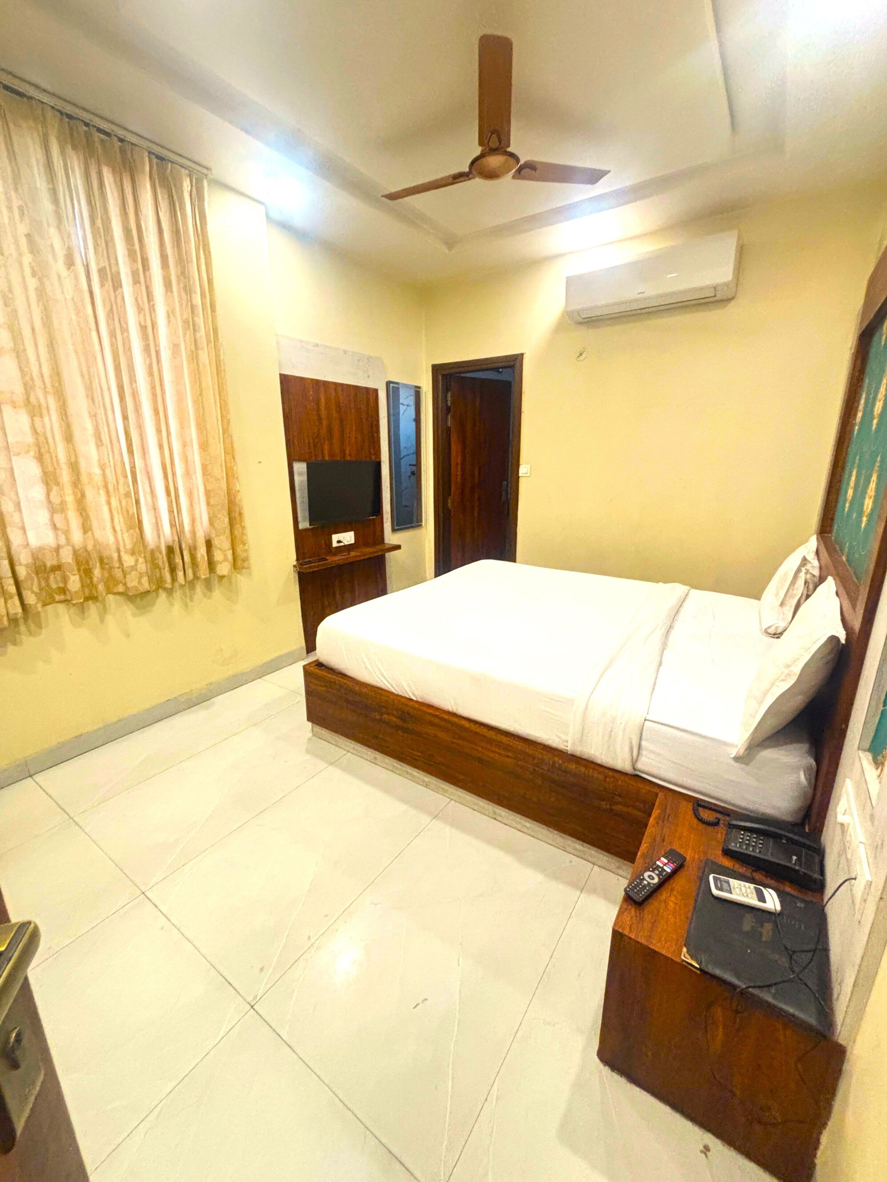 A hotel room with a bed, a TV, a ceiling fan, and a bedside table with a phone and remote controls.