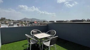 Una terraza en la azotea con mesa y sillas, vista a edificios y montañas en la distancia, y piso de césped artificial.