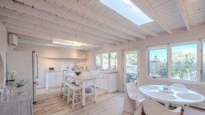 A modern kitchen with white cabinets, a dining area with a round table and white chairs, and large windows offering a view of the outdoors.
