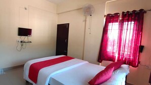 A bedroom with a bed, a TV mounted on the wall, a door, and a window with red curtains. - Shanthala Residency (Virajpet)