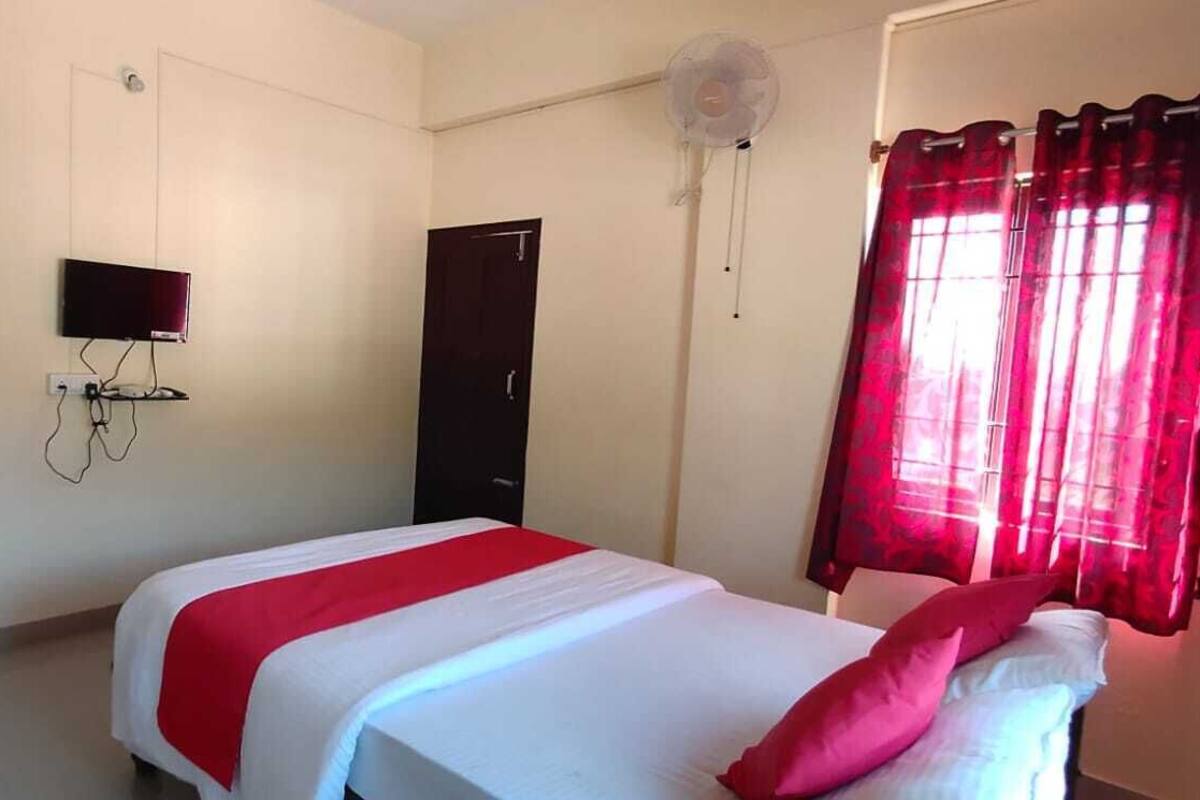 A bedroom with a bed, a TV mounted on the wall, a door, and a window with red curtains.