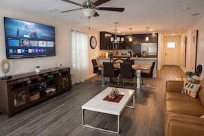 A modern living room with a flat-screen TV, a dining area with a table and chairs, a kitchen with dark cabinets and stainless steel appliances, and a ceiling fan.