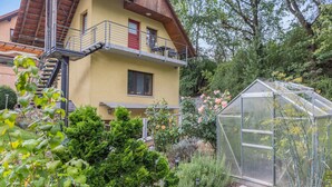 Una casa de dos pisos con un balcón, un invernadero y un jardín lleno de plantas y flores.