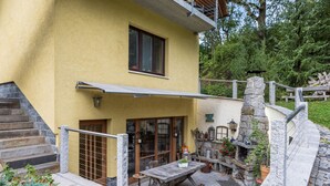 Edificio de dos pisos con fachada amarilla, una escalera de madera, un balcón con barandilla metálica y un muro de piedra con chimenea.